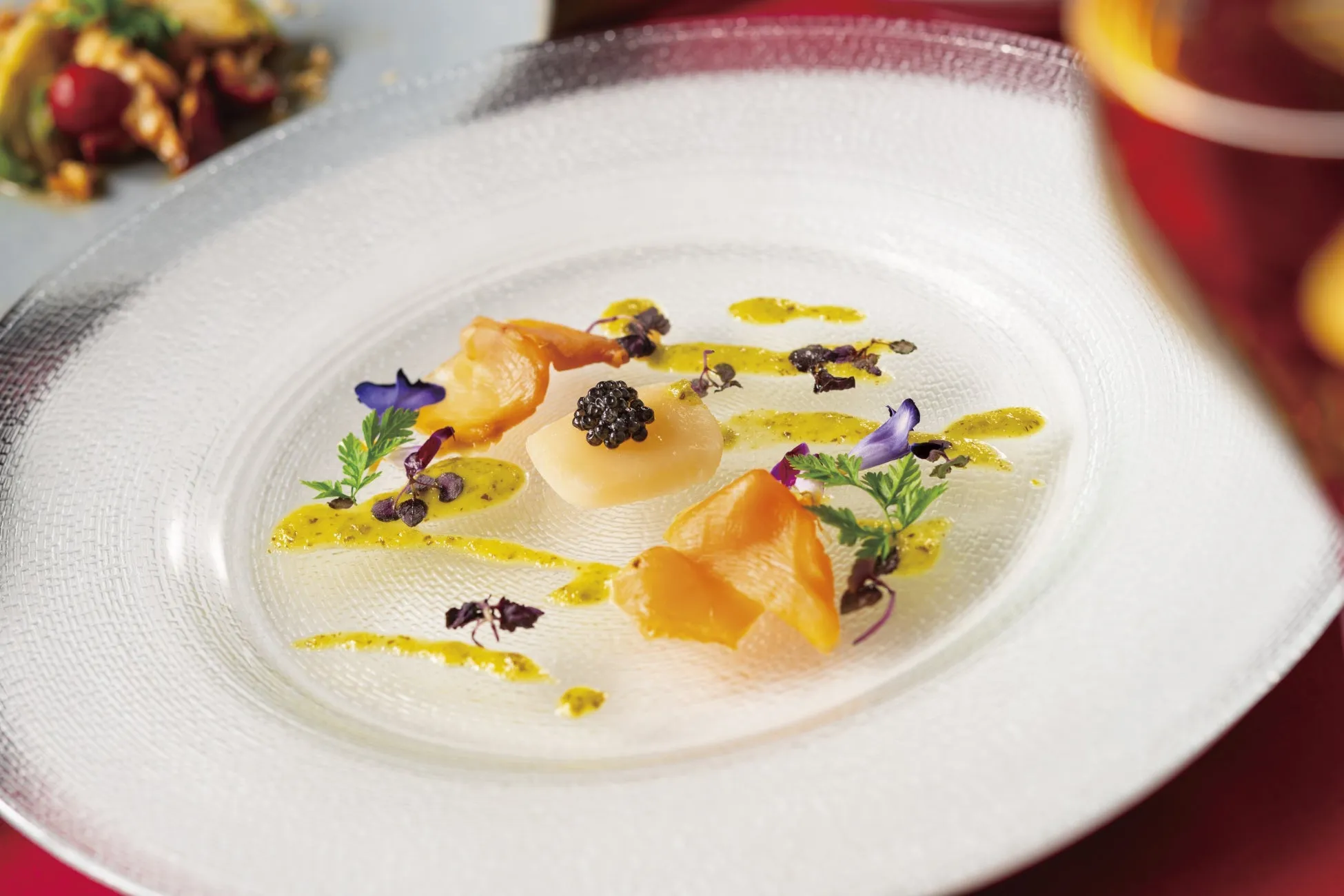 透明なガラスの皿に盛り付けられた芸術的な料理。中央にはキャビアが乗った白い食材があり、周りを黄色いソースと紫の花、ハーブが飾っている。