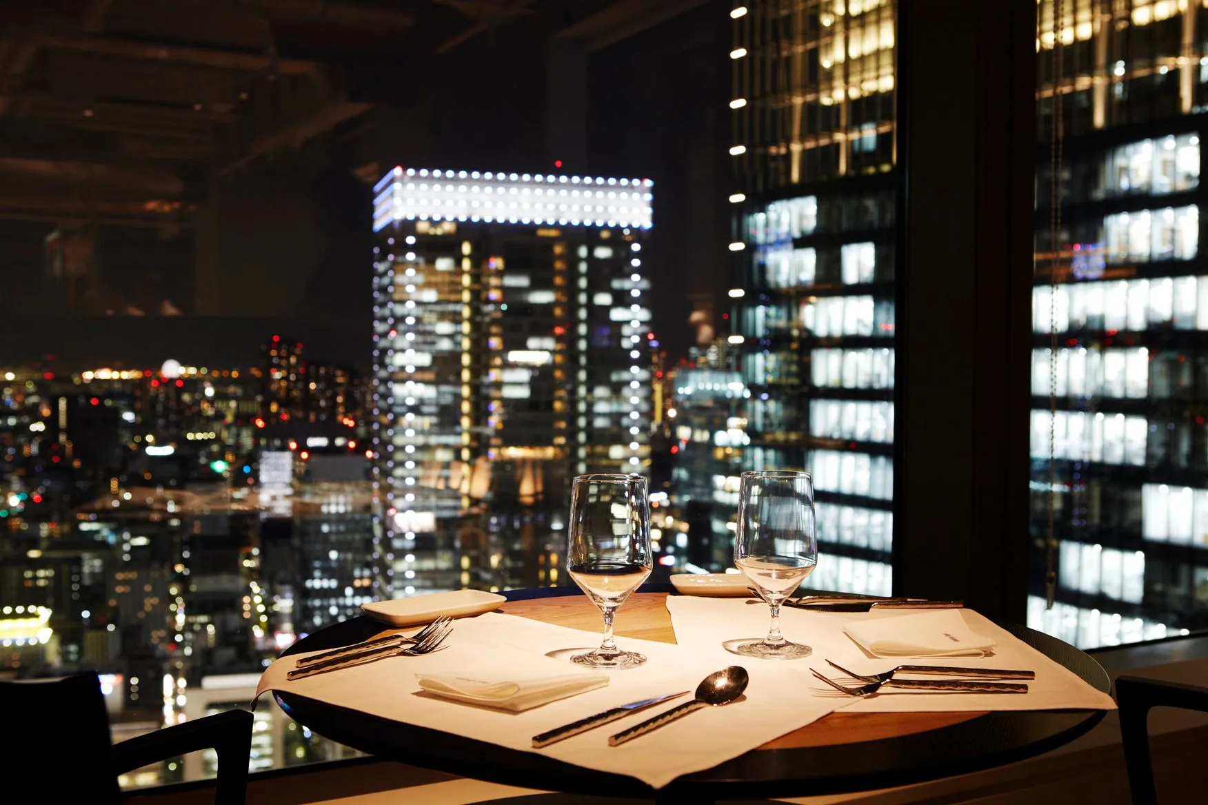 夜景を背景に、レストランのテーブルがセットされている写真。グラス、カトラリー、ナプキンが配置され、窓からはきらびやかな都市の明かりが見える。